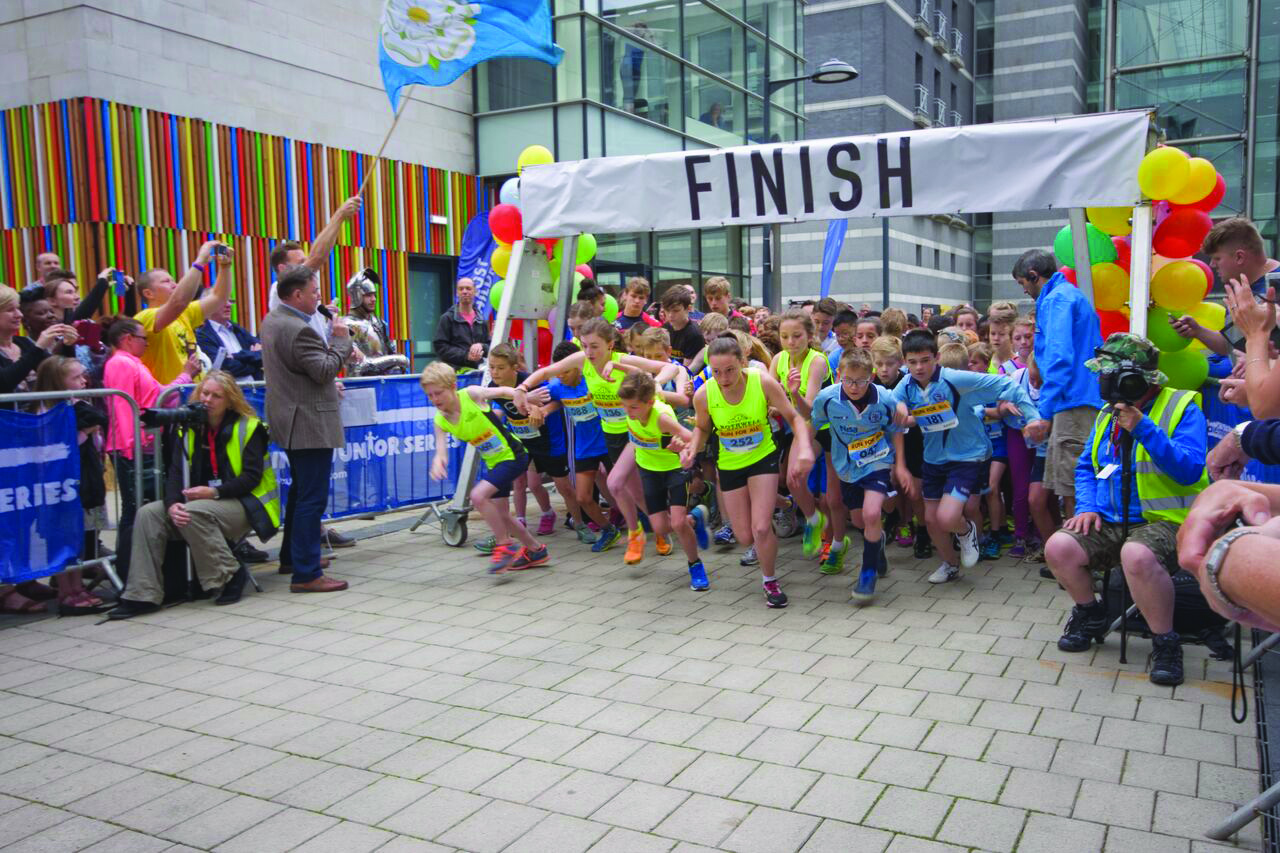 Children of all ages running in a race at Leeds Dock with a Yorkshire flag being flown by a spectator